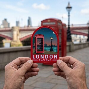 May include: A red patch featuring a London telephone booth and a lamppost against a night sky backdrop. The patch reads "LONDON ENGLAND" in white letters. The patch is held in front of a blurred background of a bridge and a red telephone booth.