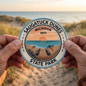 May include: A round embroidered patch featuring a scenic view of Saugatuck Dunes State Park in Michigan. The design includes a sunset over the water, dunes, and the text "SAUGATUCK DUNES MICHIGAN 1977 STATE PARK" in a circular layout. The patch is held by two hands.
