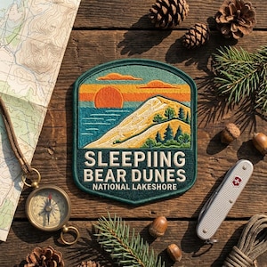 May include: A rectangular embroidered patch with rounded top corners featuring a landscape of a sun, water, and dunes. The text reads SLEEPIING BEAR DUNES NATIONAL LAKESHORE. The patch is dark green with orange, yellow, and blue thread details.