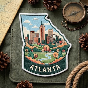 May include: An embroidered patch shaped like the state of Georgia featuring a skyline of Atlanta with buildings, a Ferris wheel, trees, and a winding river. The word ATLANTA is stitched in white at the bottom against a dark green background.