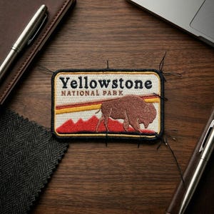 May include: A rectangular embroidered patch featuring the text Yellowstone National Park in black lettering. The design includes a brown bison silhouette, a horizontal yellow stripe, and red mountain shapes against a cream-colored background.
