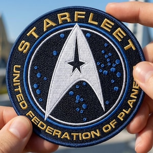 May include: Circular patch with the Starfleet insignia from Star Trek. The patch features a white arrowhead design with a star, surrounded by the words "STARFLEET" and "UNITED FEDERATION OF PLANETS" in gold lettering on a navy blue background.