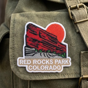 May include: An embroidered patch featuring a red rock formation against a bright orange sun, with green accents at the base. The patch has a white border and a tan banner at the bottom with the white text RED ROCKS PARK COLORADO.