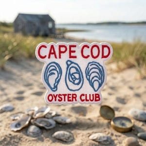 May include: A white embroidered patch with red text that reads CAPE COD OYSTER CLUB. The patch features three blue embroidered oyster shell illustrations centered between the two lines of text, with a scalloped edge design.