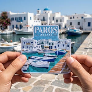 May include: A rectangular embroidered patch featuring a scenic harbor view of Paros, Greece. The design includes white buildings, blue and white boats on turquoise water, and a brown stone pier. The text PAROS GREECE is stitched in white.