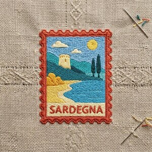 May include: A rectangular embroidered patch shaped like a postage stamp with a scalloped rust-colored border. The design depicts a coastal landscape with a yellow sun, blue sky, white clouds, green hills, a stone tower, and the text SARDEGNA.