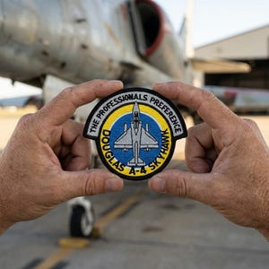 May include: A circular patch with the text "THE PROFESSIONALS PREFERENCE DOUGLAS A-4 SKYHAWK" encircling a blue and yellow emblem of an aircraft. The patch is held in front of an aircraft.