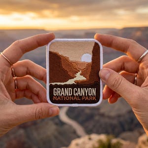 May include: A rectangular patch featuring a scenic view of the Grand Canyon at sunset. The patch has a white border and includes the text "GRAND CANYON NATIONAL PARK" in white lettering. The image depicts the canyon's layered rock formations and a river.