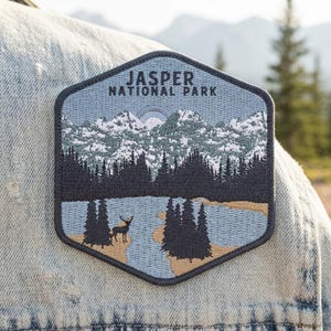 May include: A hexagonal embroidered patch featuring a landscape of Jasper National Park. The design includes snow-capped mountains, a dark forest, a blue lake, and a deer standing on the shore, all stitched in shades of blue, white, and tan.