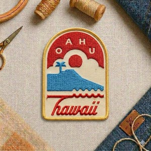 May include: Embroidered patch with a Hawaii design. The patch features a red arch with the letters "AHU", a red sun, a blue island, and the word "Hawaii" in red script. The patch has a yellow border.