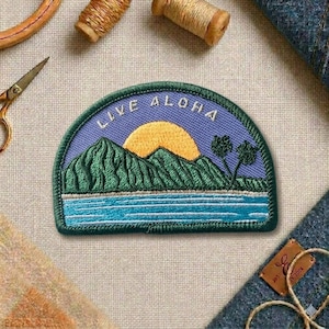 May include: Embroidered patch with a landscape design featuring mountains, a setting sun, and palm trees. The patch has the words "LIVE ALOHA" in white lettering on a blue background. The patch is surrounded by a green border.