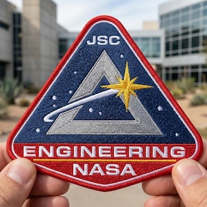 May include: Embroidered patch with a triangular design featuring a starburst, a stylized comet, and the text "JSC" at the top. The patch has a red border and the words "ENGINEERING NASA" at the bottom. The background is blue.
