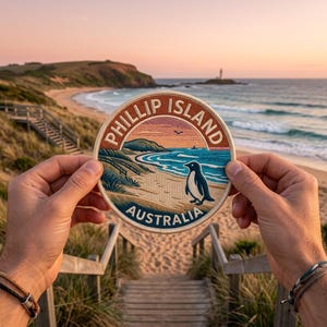 May include: A circular embroidered patch featuring a coastal landscape with a penguin on a sandy beach, blue ocean waves, and grassy dunes. The text PHILLIP ISLAND and AUSTRALIA is stitched in white lettering along the top and bottom borders.