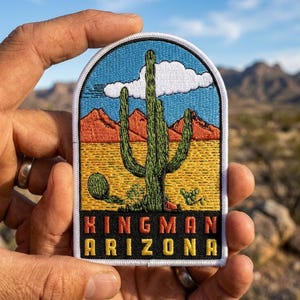 May include: An embroidered patch with an arched top featuring a desert landscape with a tall green saguaro cactus, orange mountains, a white cloud, and a blue sky. The text KINGMAN ARIZONA is stitched in orange and yellow at the bottom.
