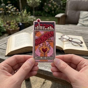 May include: A rectangular embroidered patch featuring a matchbox design with the text SLOW BURN MATCHES and MADE FOR READERS. The design includes a row of matches at the top, a flame graphic with a heart center, and a pink, orange, and cream color palette.
