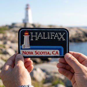 May include: A black patch with the word "HALIFAX" in white, and a lighthouse illustration. Below, it reads "NOVA SCOTIA, CA" in red and white. The patch has a blue and white striped border and a red top. The background shows a lighthouse and the ocean.