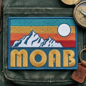 May include: A rectangular embroidered patch featuring a mountain range in white against a sunset background of teal, yellow, orange, and red horizontal stripes. A white circle represents the sun. The word MOAB is stitched in large yellow letters.
