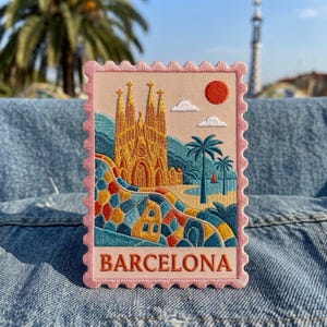 May include: A rectangular embroidered patch shaped like a postage stamp with a pink scalloped border. It features a golden cathedral, a colorful mosaic wall, palm trees, a red sun, and white clouds. The word BARCELONA is stitched in orange.