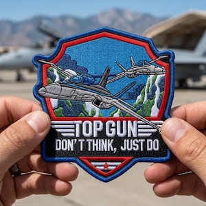 May include: Embroidered patch featuring two fighter jets in flight over a mountain range, with the text "TOP GUN DON'T THINK, JUST DO." The patch has a blue, red, and white color scheme, with a shield-like shape.