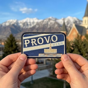 May include: A rectangular patch with the word "PROVO" in white block letters above a depiction of a building and the word "UTAH". The patch has a blue background with white and tan accents. The patch is held in front of a mountain range.