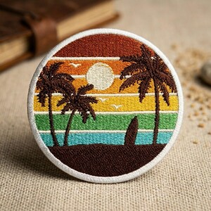 May include: Embroidered patch featuring a sunset beach scene. The design includes palm trees, a surfboard, and a sun, with a color palette of orange, yellow, green, and blue. The patch is round with a white border.