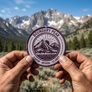 May include: A round, embroidered patch with the text "Boundary Peak" and mountain graphics. The patch also includes the text "4,005M - Nevada - 13,140FT". The patch is held in front of a mountain range.