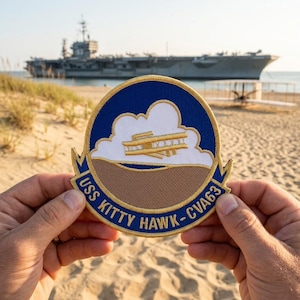 May include: A round patch with a blue border and the text "USS KITTY HAWK - CVA63". The patch features a cloud design with a gold and white airplane. The background shows an aircraft carrier on a beach.