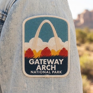 May include: An embroidered patch on a light blue denim jacket featuring the Gateway Arch in cream against a blue sky and white clouds, with red and yellow hills below. The text reads GATEWAY ARCH NATIONAL PARK in white letters.