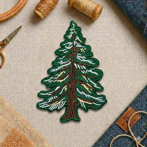 May include: Embroidered patch in the shape of a green pine tree with brown trunk and white accents. The patch is on a neutral fabric surface, with sewing supplies in the background.