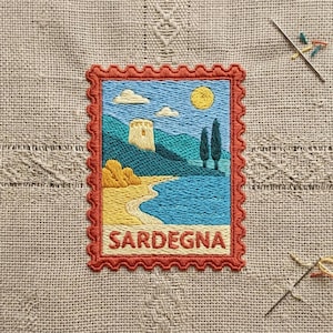 May include: A rectangular embroidered patch shaped like a postage stamp featuring a coastal landscape with a stone tower, yellow sun, blue sea, and sandy beach. The word SARDEGNA is stitched in red at the bottom, framed by a scalloped red border.
