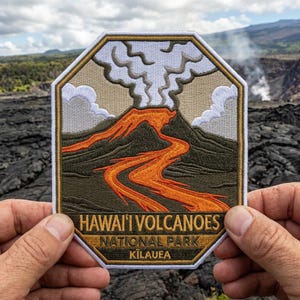 May include: Embroidered patch featuring a volcano with orange lava flowing down a dark green mountain. The patch has the text "HAWAI'I VOLCANOES NATIONAL PARK KILAUEA" in a gold border. The background shows a volcanic landscape.