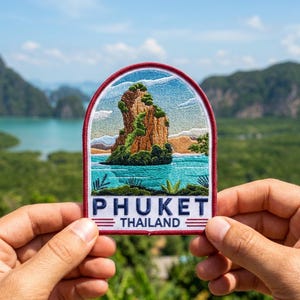 May include: An embroidered patch with a rounded top edge featuring a landscape of a rocky island in turquoise water. The design includes green vegetation, a blue sky, and the text PHUKET THAILAND in dark blue letters with red horizontal lines.