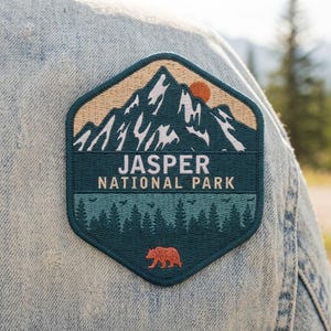 May include: A hexagonal embroidered patch featuring a mountain range with a sun, a forest of pine trees, and a bear silhouette. The text reads JASPER NATIONAL PARK in white capital letters against a dark teal background on the patch.