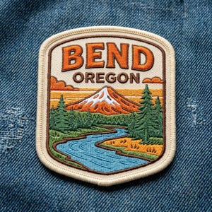 May include: A rectangular embroidered patch with rounded corners featuring a landscape scene of a snow-capped mountain, a winding blue river, and green pine trees. The text BEND OREGON is stitched in orange and brown letters at the top.
