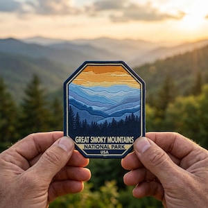May include: A rectangular patch featuring a mountain landscape with a blue and orange sky. The patch reads "Great Smoky Mountains National Park USA" in white text. The patch is held between two hands, with a blurred mountain backdrop.