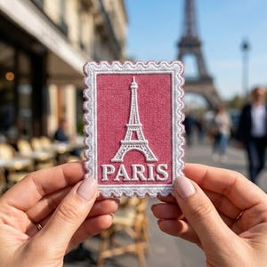 May include: A rectangular embroidered patch shaped like a postage stamp with a scalloped white border. The center features a white stitched Eiffel Tower graphic above the word PARIS in white capital letters on a solid dusty rose pink background.