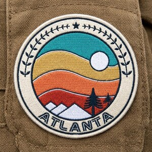 May include: A circular embroidered patch featuring a landscape with teal, yellow, orange, and red wavy bands, a white sun, white mountain peaks, and dark blue pine trees. The border is cream with a leaf pattern and the text ATLANTA.