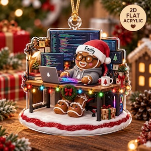 May include: A 2D flat acrylic Christmas ornament featuring a gingerbread person wearing a Santa hat with the name Emily, sitting at a desk with a laptop, computer monitors displaying code, a mug, and colorful string lights.