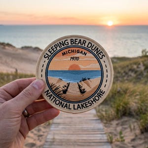 May include: A round patch with the text "Sleeping Bear Dunes Michigan 1970 National Lakeshore" features a sunset over a lake and dunes. The patch is beige with a blue, orange, and brown design. The patch is held in front of a wooden walkway.
