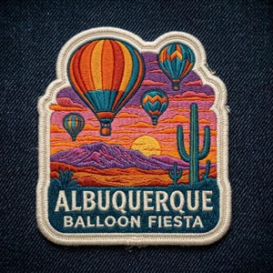 May include: An embroidered patch featuring a desert landscape with four hot air balloons in orange, yellow, and blue, a green saguaro cactus, and a yellow sun over purple mountains. The text reads ALBUQUERQUE BALLOON FIESTA in white letters.