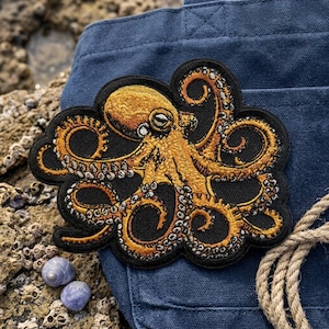 May include: Embroidered octopus patch with gold and black thread. The octopus is detailed with multiple tentacles and a central head. The patch is on a black background with a blue fabric in the background. A rope and seashells are also visible.