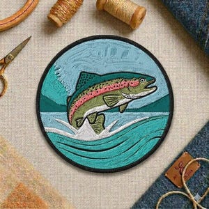 May include: Embroidered patch featuring a jumping rainbow trout. The patch has a circular shape with a black border. The trout is green, pink, and red, set against a blue and teal water and sky background. The patch is likely for clothing or accessories.