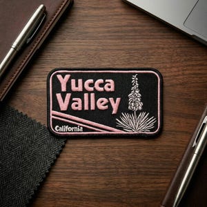 May include: A rectangular embroidered patch with a black background and a light pink border. The text reads Yucca Valley in large pink letters with California in smaller text below. A stylized pink yucca plant graphic is on the right side.
