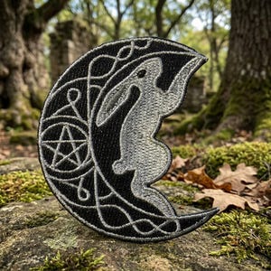 May include: Black and white embroidered patch in the shape of a crescent moon. The patch features a stylized rabbit climbing the moon, with a pentagram and decorative swirls. The patch is likely for clothing or accessories.