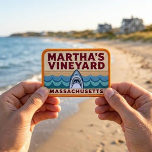 May include: A rectangular patch with the words "MARTHA'S VINEYARD MASSACHUSETTS" in maroon and teal. The patch features a shark graphic with an open mouth, waves, and a tan background. The patch is held up in front of a beach scene.