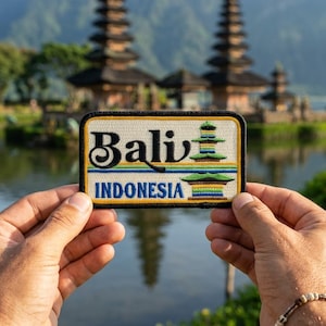 May include: A rectangular patch with the word "Bali" in a stylized font and "Indonesia" below. The patch features a colorful pagoda design and a black border. The patch is held in front of a lake and temple background.