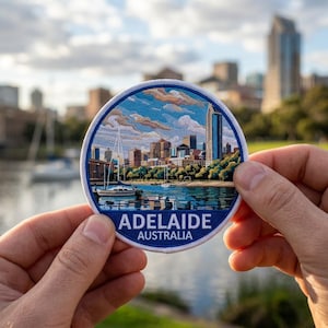 May include: A round embroidered patch featuring a detailed cityscape of Adelaide, Australia, with a blue and white border. The patch displays the city's skyline, a river, and the text "ADELAIDE AUSTRALIA".