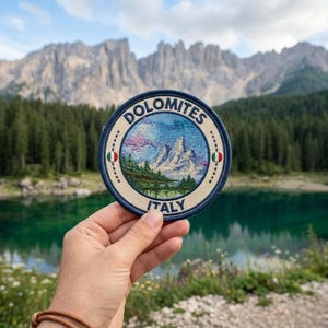 May include: A round embroidered patch featuring the word "Dolomites" and the word "Italy". The patch has a blue border and depicts a mountain landscape with a lake. The patch is held in front of a scenic background of mountains and water.