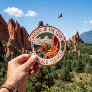 May include: A circular patch with the text "GARDEN OF THE GODS COLORADO SPRINGS" in a tan border. The patch features a landscape scene with red rock formations, green trees, and a blue sky. A person's hand holds the patch in front of a similar background.