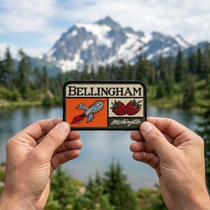 May include: A rectangular patch with the word "Bellingham" at the top. The patch features a rocket ship and raspberries, with "Washington" below. The patch is held in front of a mountain and lake.